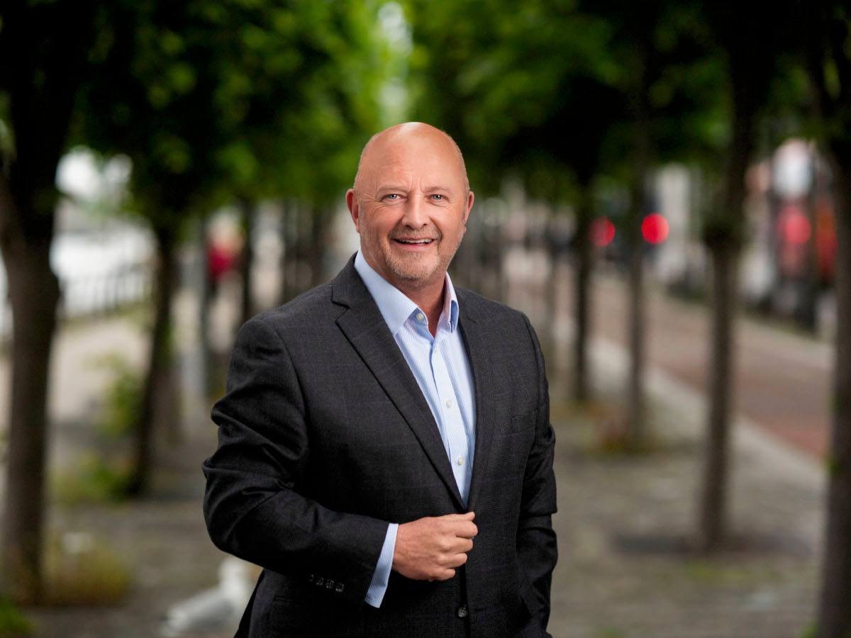 A bald, middle-aged white man in a suit posing in front of blurred trees.