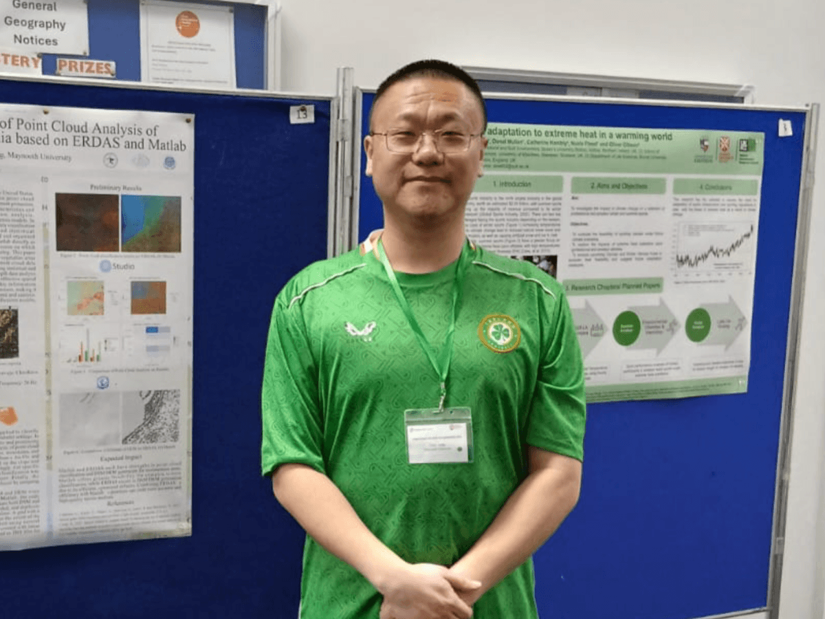 A man wearing a green T shirt and glasses, standing with his hands clasped. He is standing in front of a blue pin board.