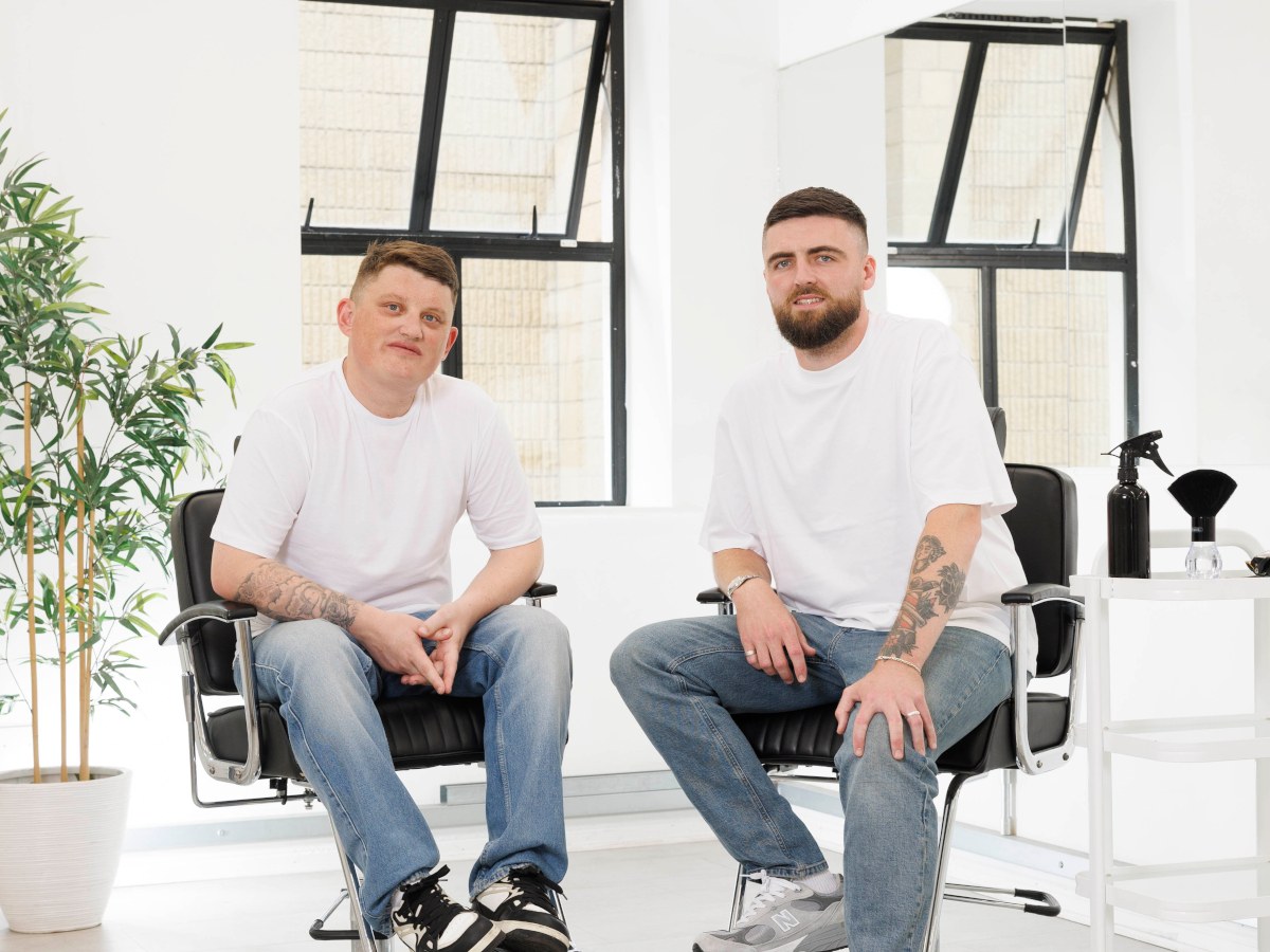 Two young men in white tees and jeans sit in a white salon on barber chairs.