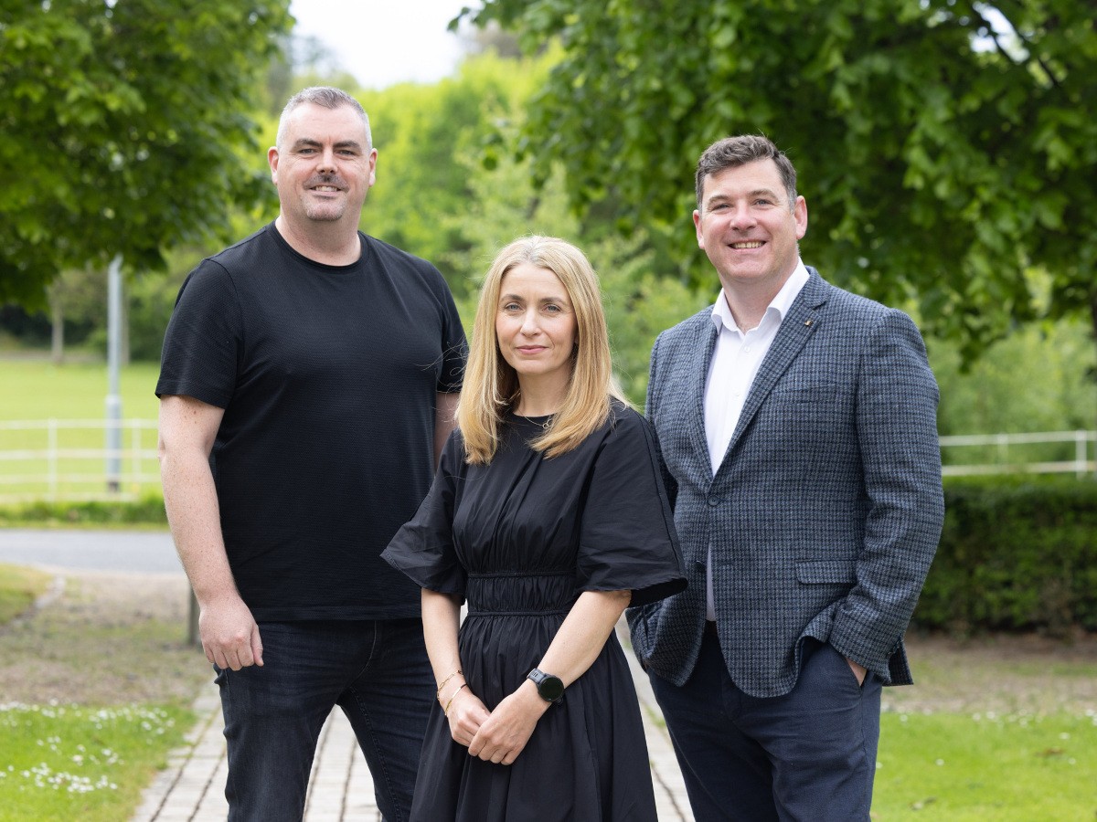 Two men and a woman in the centre standing, smiling. The three are standing in day light with greenery in the background.