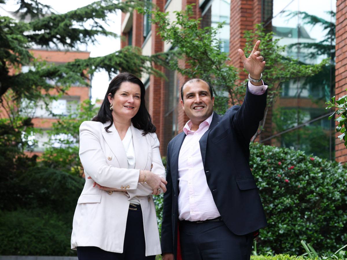 A woman with short, dark hair and a white blazer standing to the right of a man in a dark blazer and light-coloured shirt pointing towards something ahead of them.