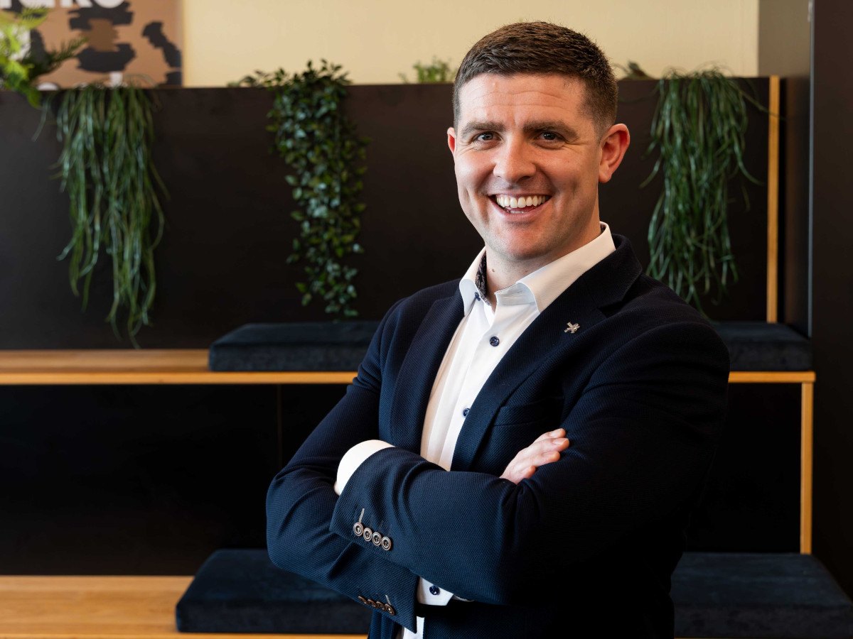 A young man, smiling, dressed in business attire posing with his arms crossed in an office.