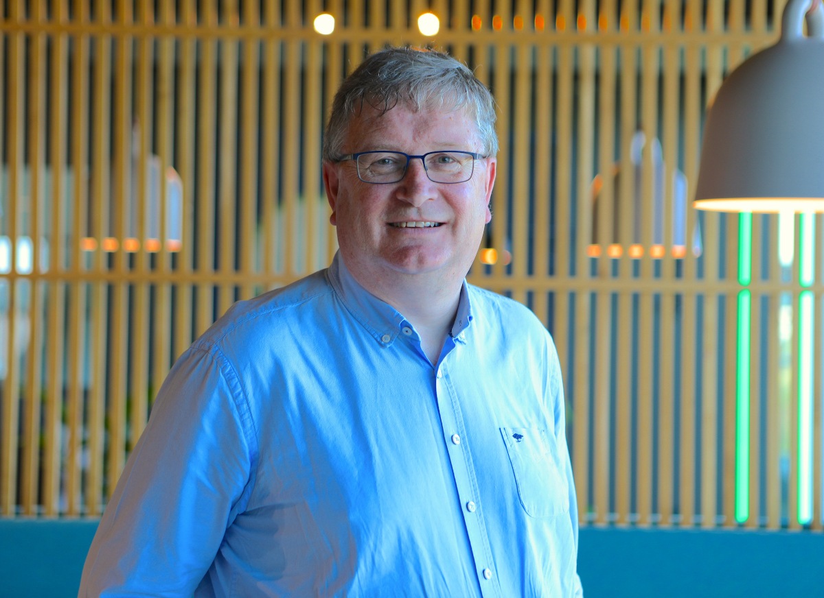 Caucasian male in open-necked blue shirt smiles to camera.