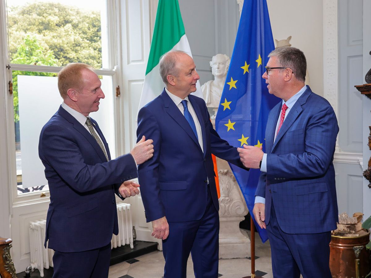 Three men in suits are interacting with each other. There is a large Irish flag and an EU flag behind them.