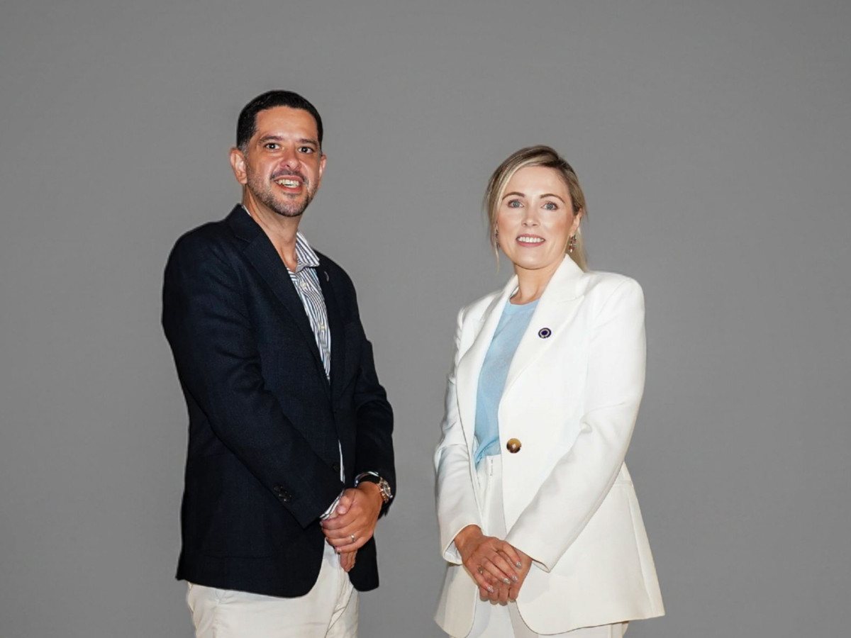 A middle-eastern man and white woman pose in front of a grey background, with their arms folded in front of themselves.