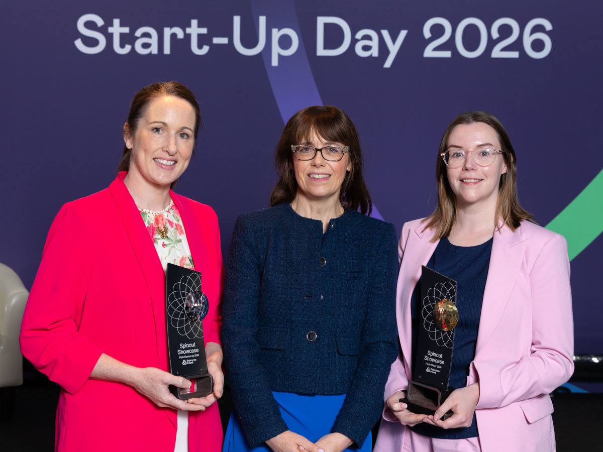 Three women in office wear, standing. The two to the sides are holding awards.