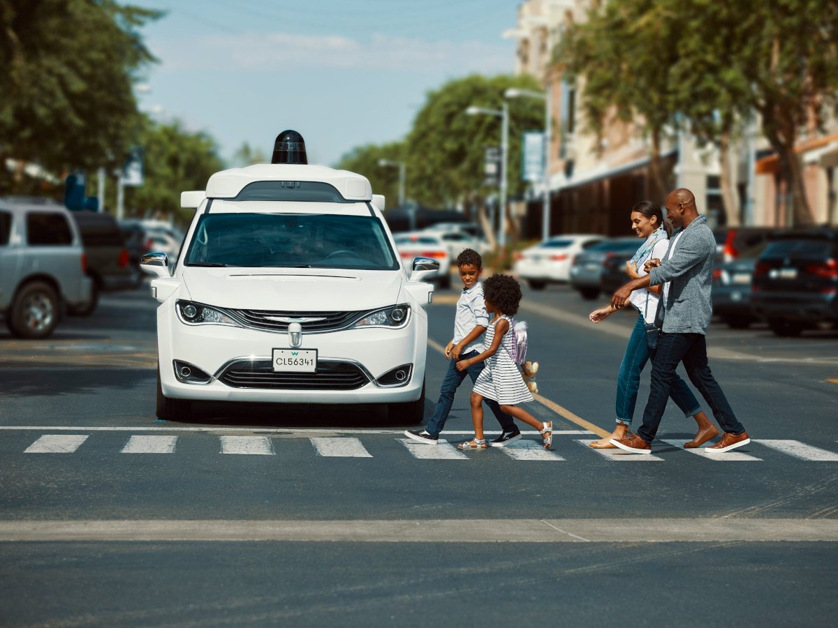 A white Waymo car stopped behind a crosswalk where a man, woman and two children are seen crossing.