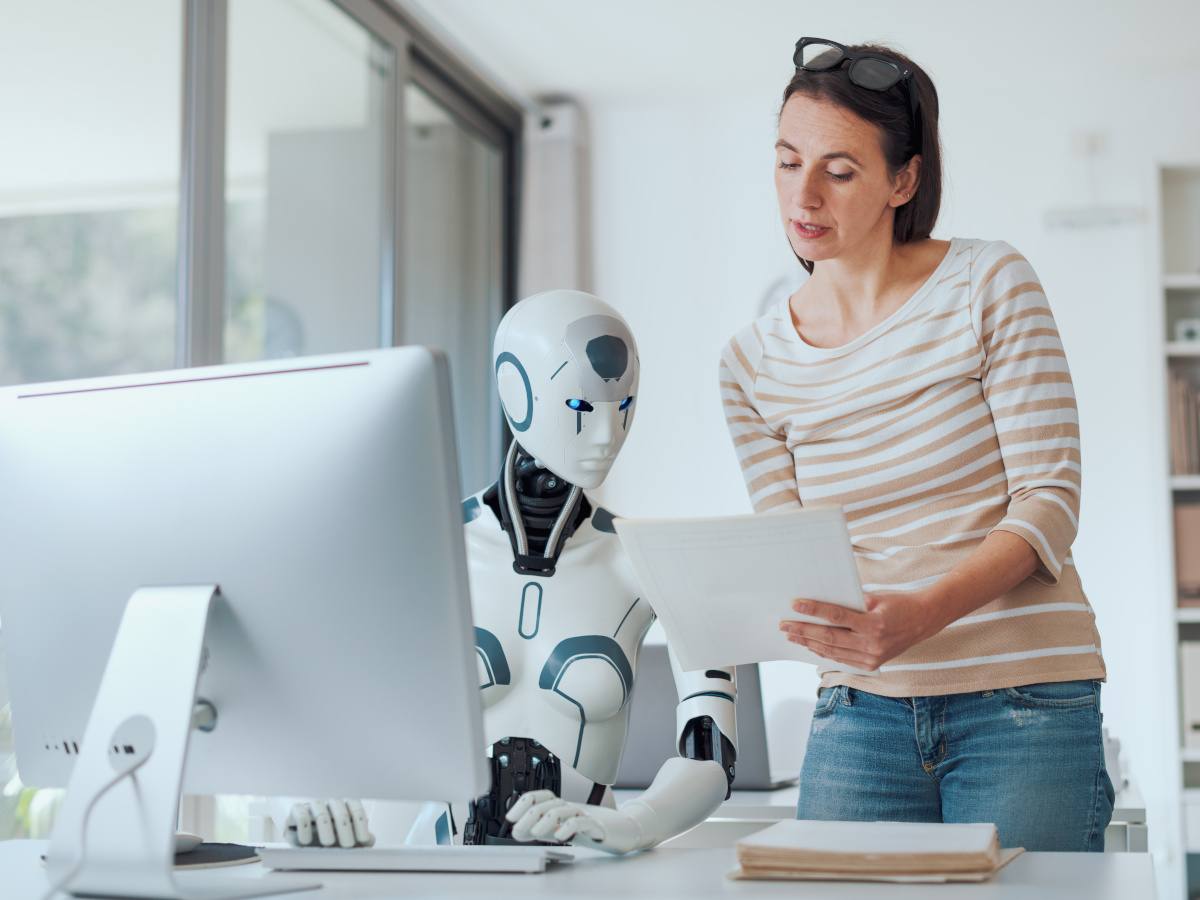 A woman in the office shows a piece of paper to an AI robot in front of a laptop.