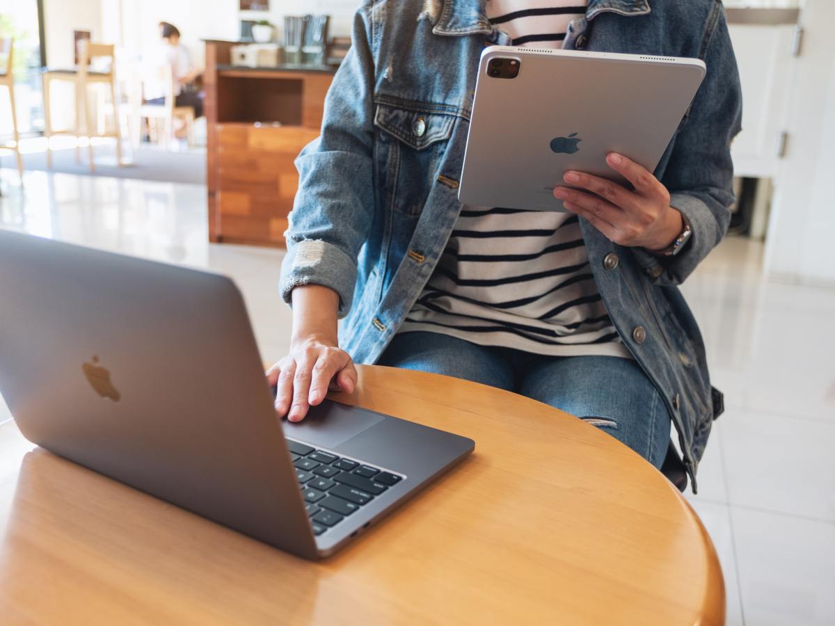 A person wearing a denim jacket holding an iPad in one hand while using a laptop with the other.