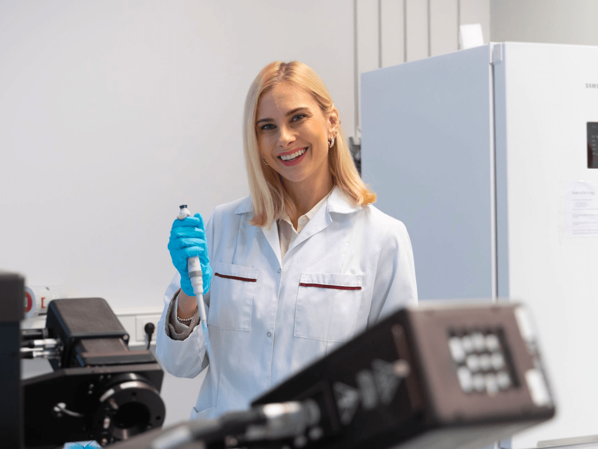 A young, white, blond woman in a lab coat and blue gloves, holding a pipette.