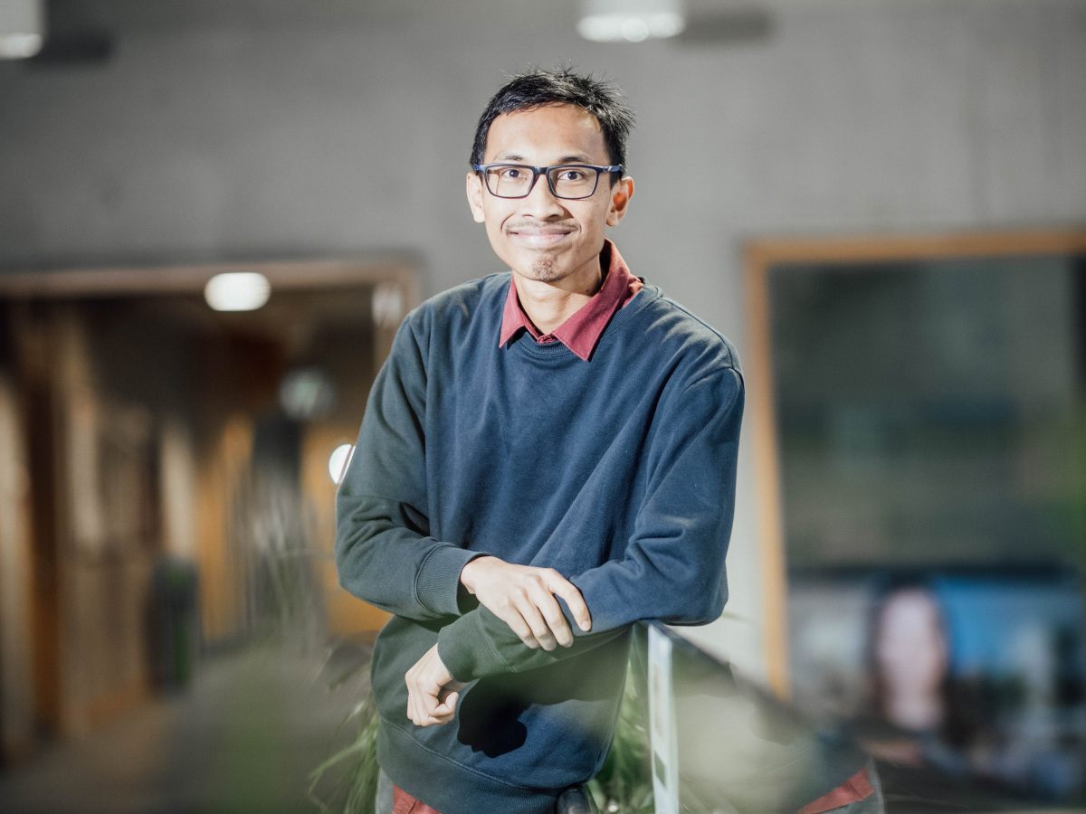 An Indonesian man with short black hair wearing glasses and a navy-gray sweatshirt over a red shirt smiles while leaning on a bannister in a hallway.