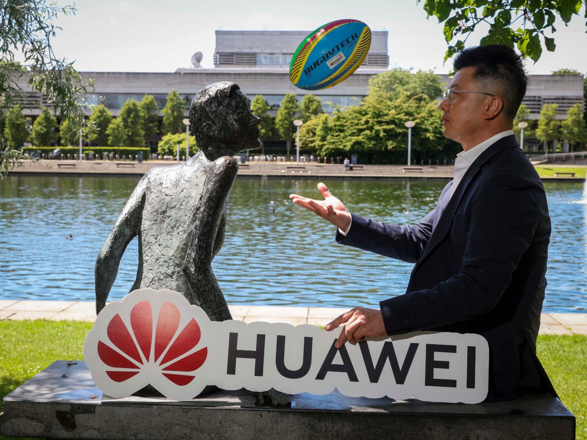 A man in a suit turned to his side with a rugby ball tossed mid way in the air. He is holding a Huawei logo in his other hand, facing the camera.