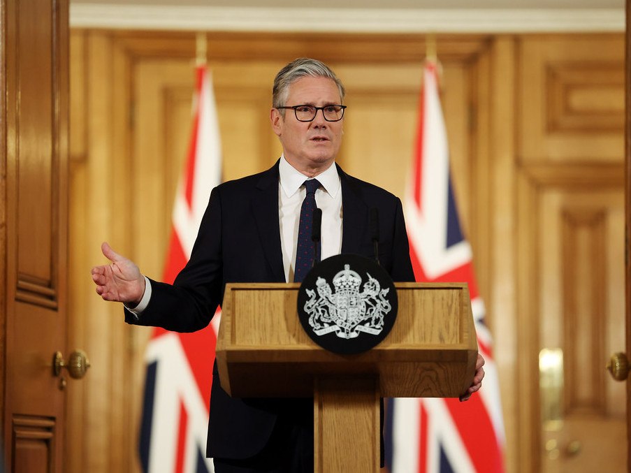 UK prime minister Keir Starmer stands at a podium with British flags behind him giving a speech wearing a dark suit and tie.
