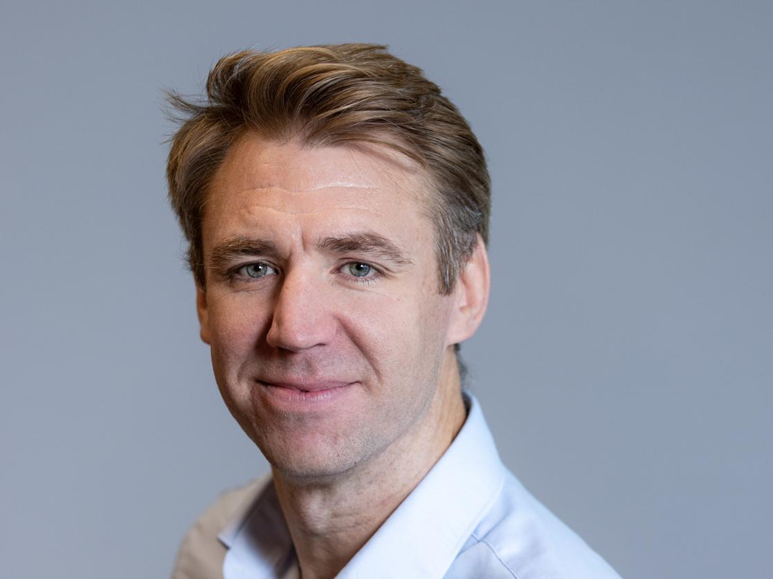 A man with short brown hair smiles while wearing a white shirt with the collar undone. He is standing in front of a grey background.