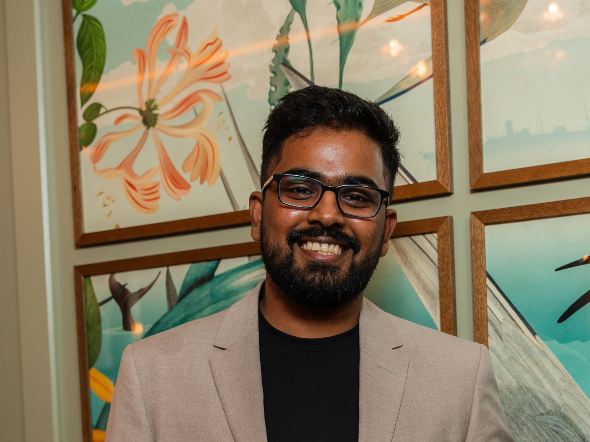 A dark-skinned South-Asian man in a black shirt and tan blazer, in front of a floral wall.