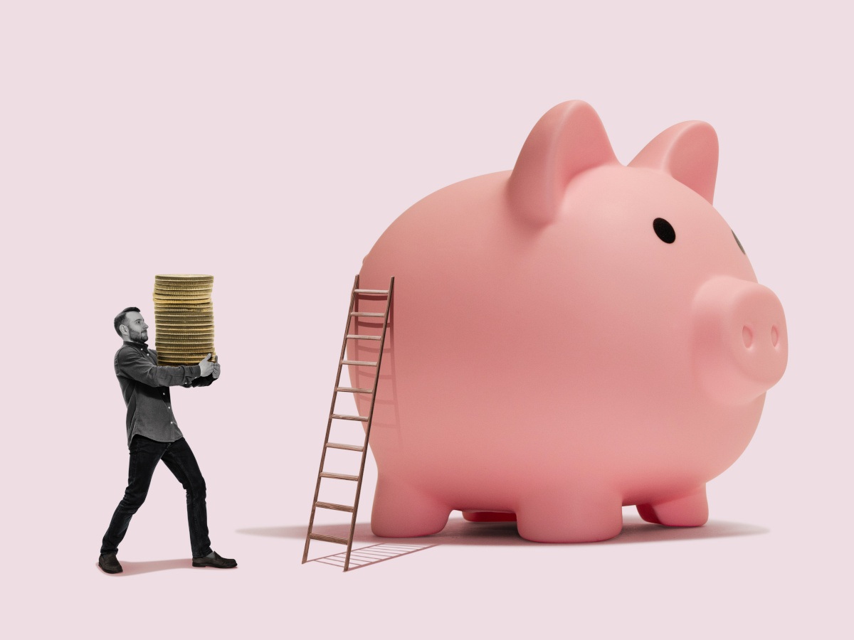 A large pink piggy bank with a ladder against it. A man carrying several plate-sized coins is walking toward the piggy bank.