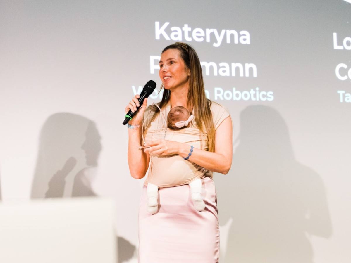 A young woman in blush, holding a microphone, with a baby strapped to her, on a stage discussing robotics.