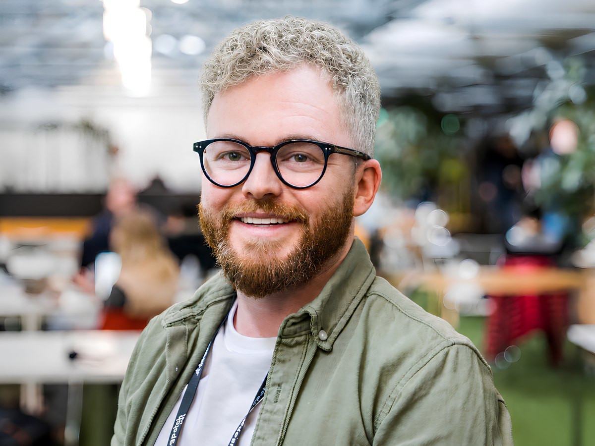 A man with short curly hair, beard and glasses smiles for the camera with a blurred office in the background.