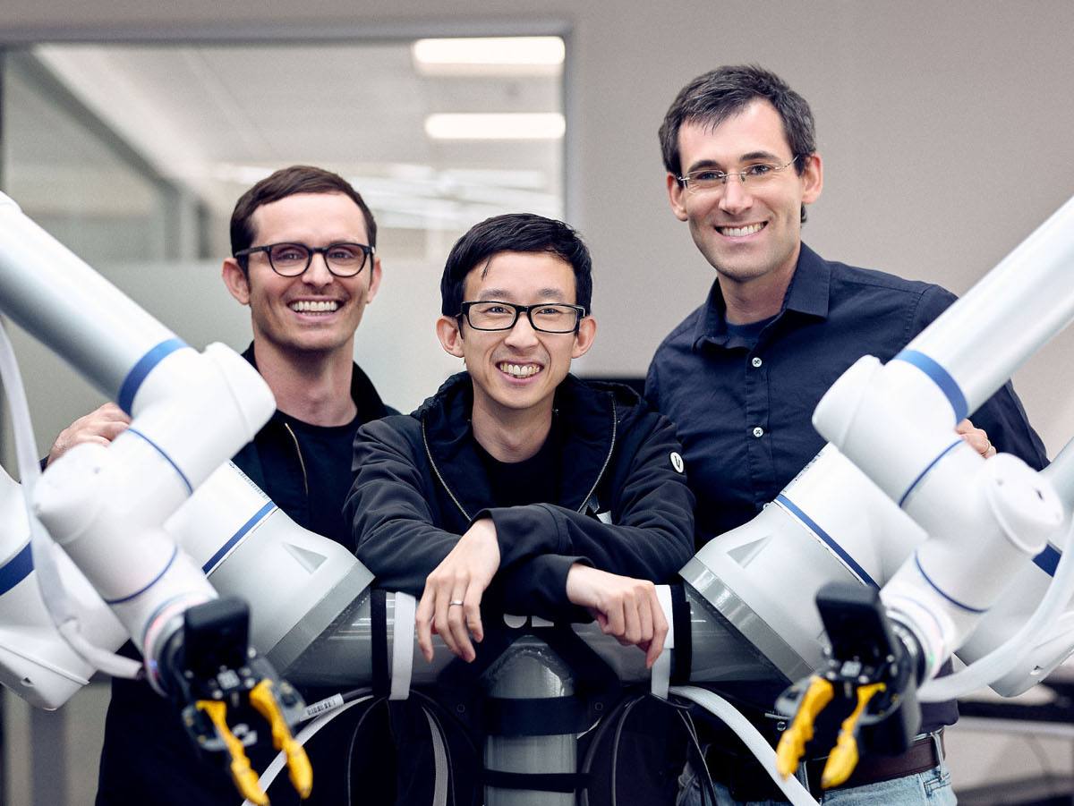 Three men smiling at the camera with a robot with two arms stretching to either side.