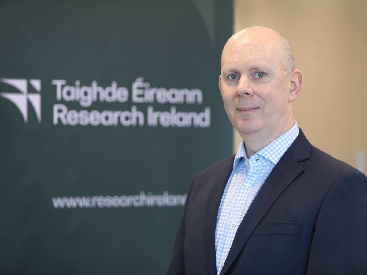 Caucasian male in suit and open necked collar stands in front of Research Ireland signage.