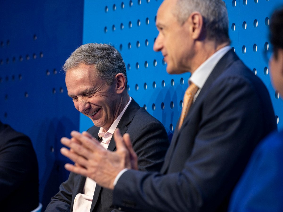 The side profile of two men in dark suits speaking at a conference.