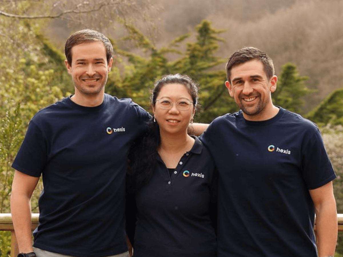 Two young white men and a young Asian woman posing outside, wearing black Hexis t-shirts.