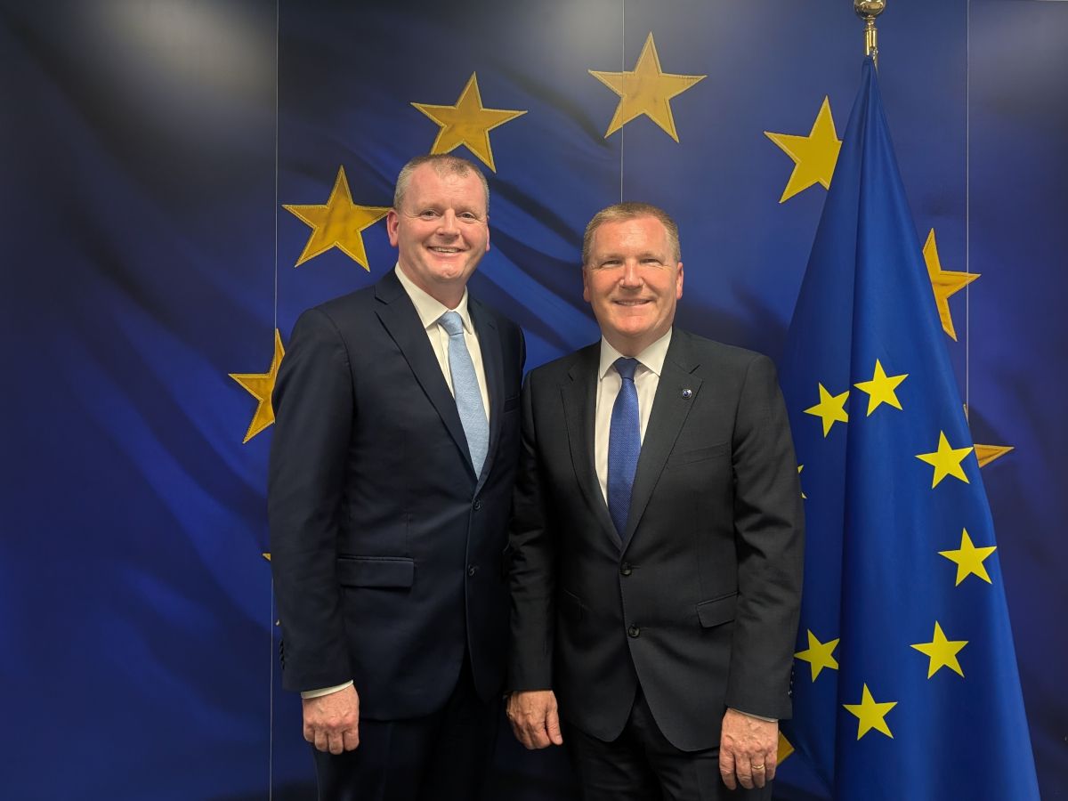 Two middle aged white men wearing suits and ties stand, smiling in front of a blue and yellow EU background.