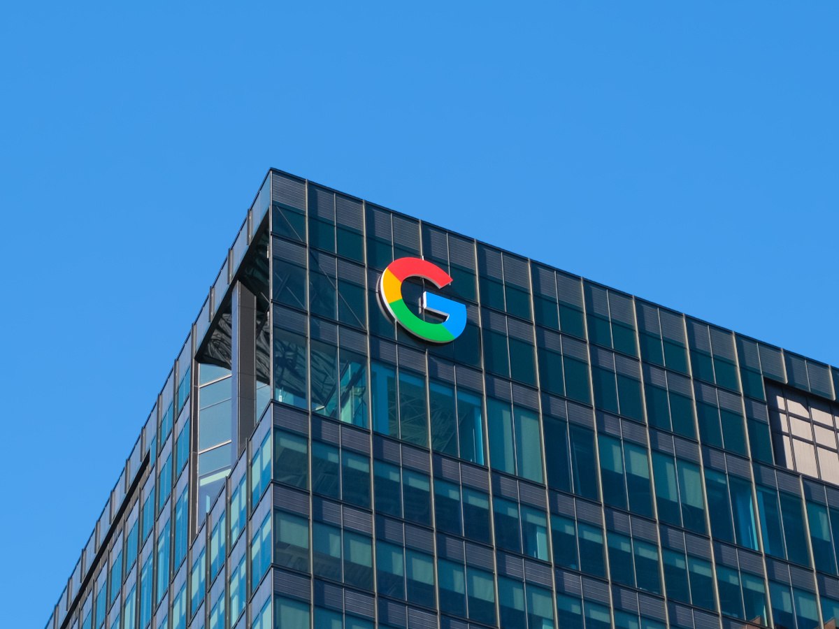 The G of the Google logo at the top of a large glass office building against a clear blue sky.