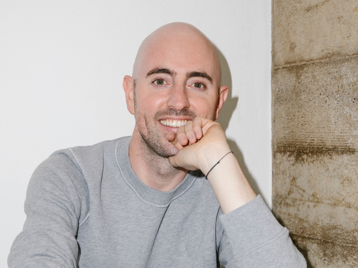 A bald white man wearing a grey jumper with a hand on his chin.