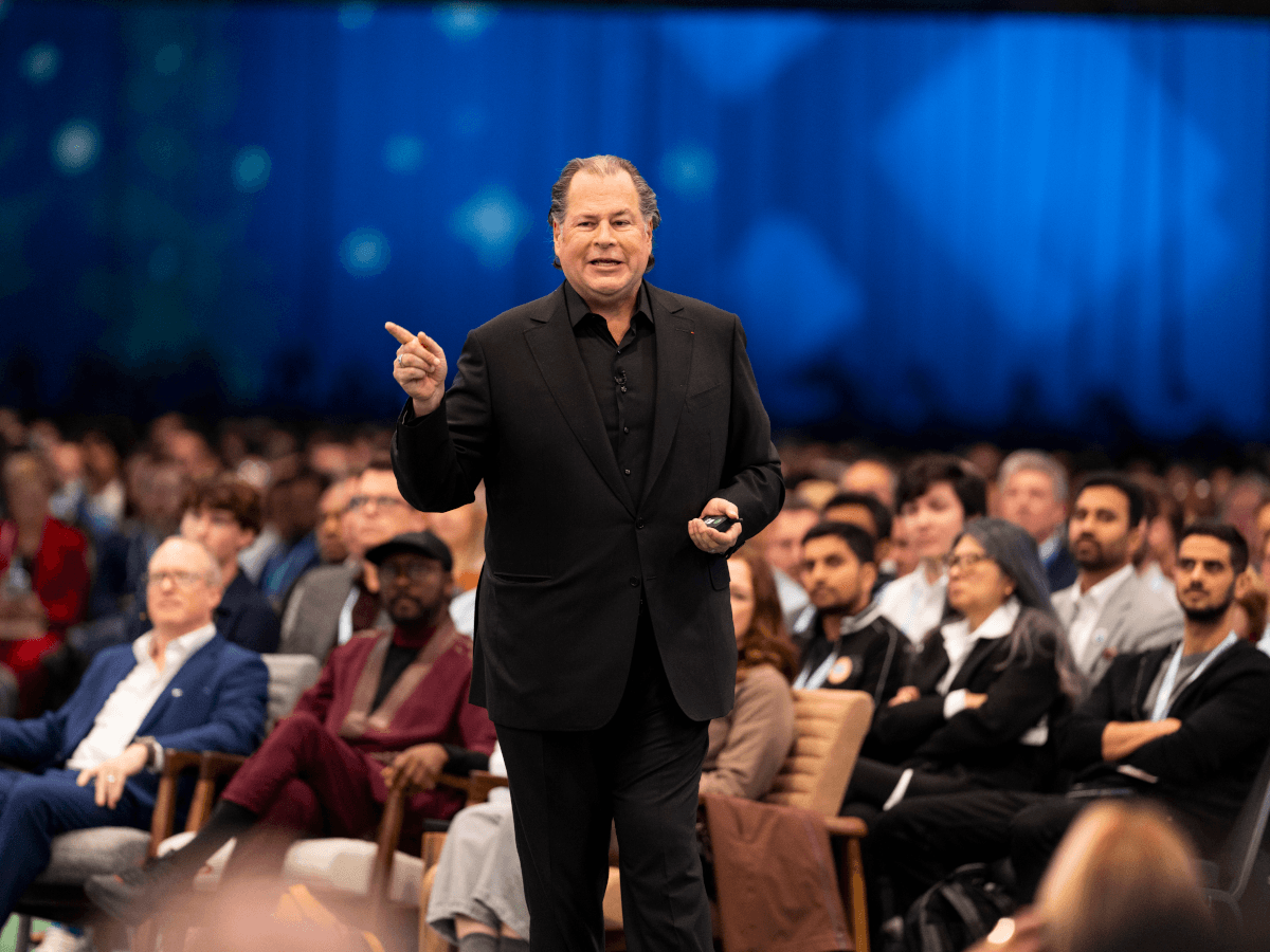 A man in a black suit standing around a crowd of people sitting, looking at him.