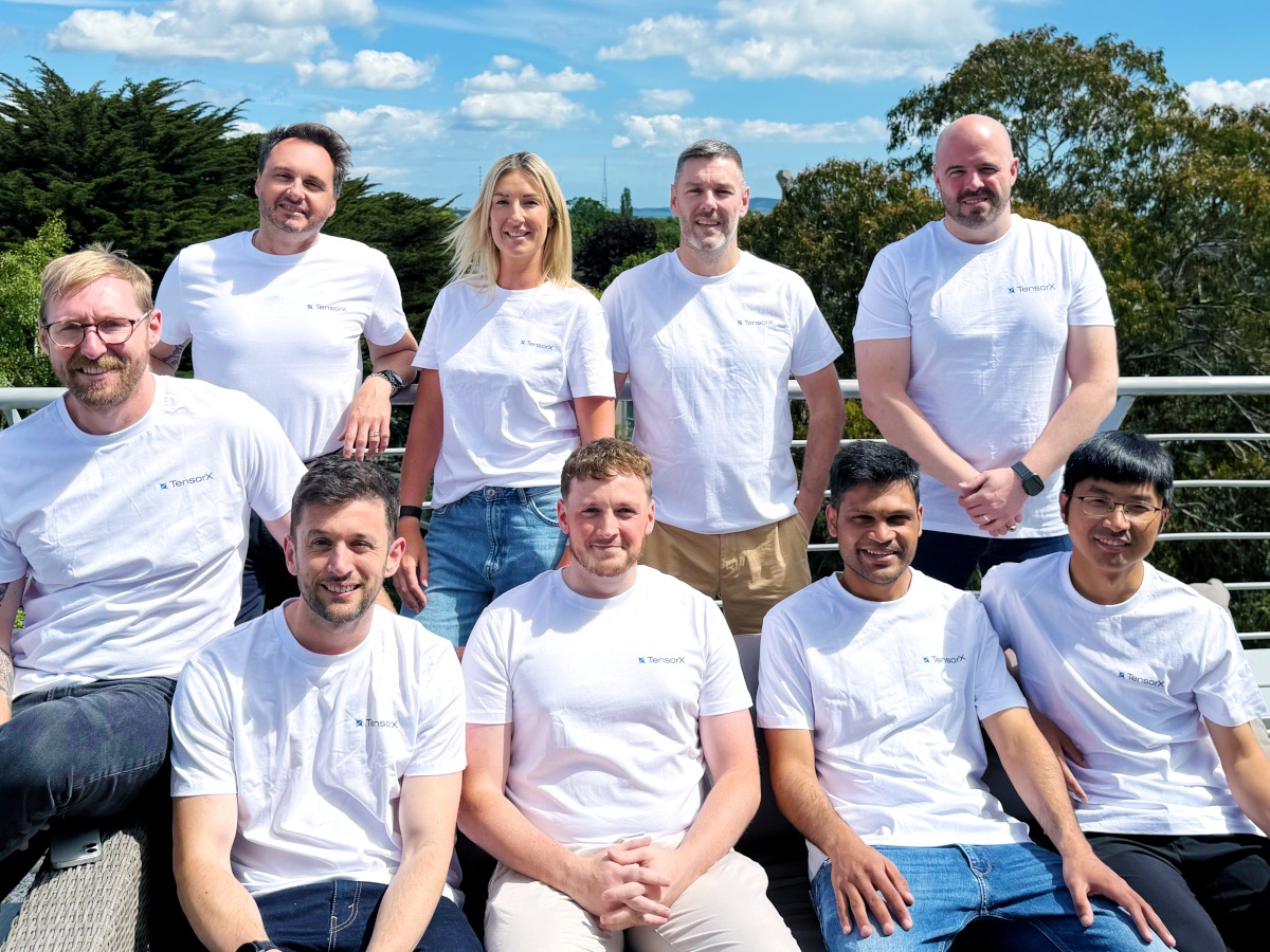 Two rows of people in white t shirts smile on a sunny day on a rooftop.