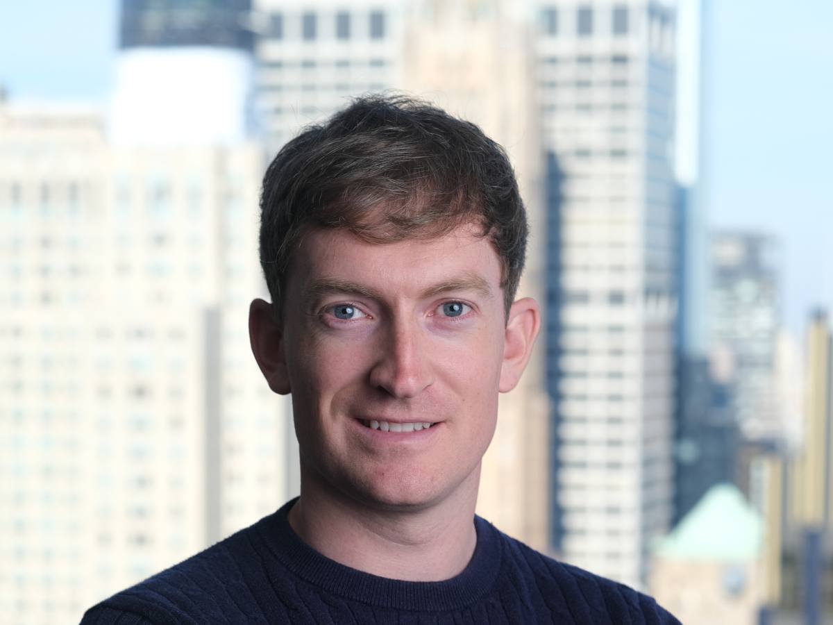 A white man with fair hair and wearing a dark sweater smiles in front of a city backdrop.