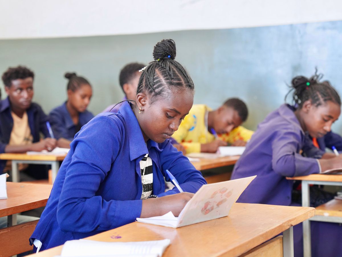 A young Ethiopian girl sits in a school classroom writing in a notebook. There are other students behind her.