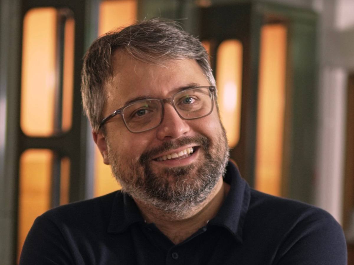 A man with greying hair and glasses smiles with his head tilted slightly to one side.