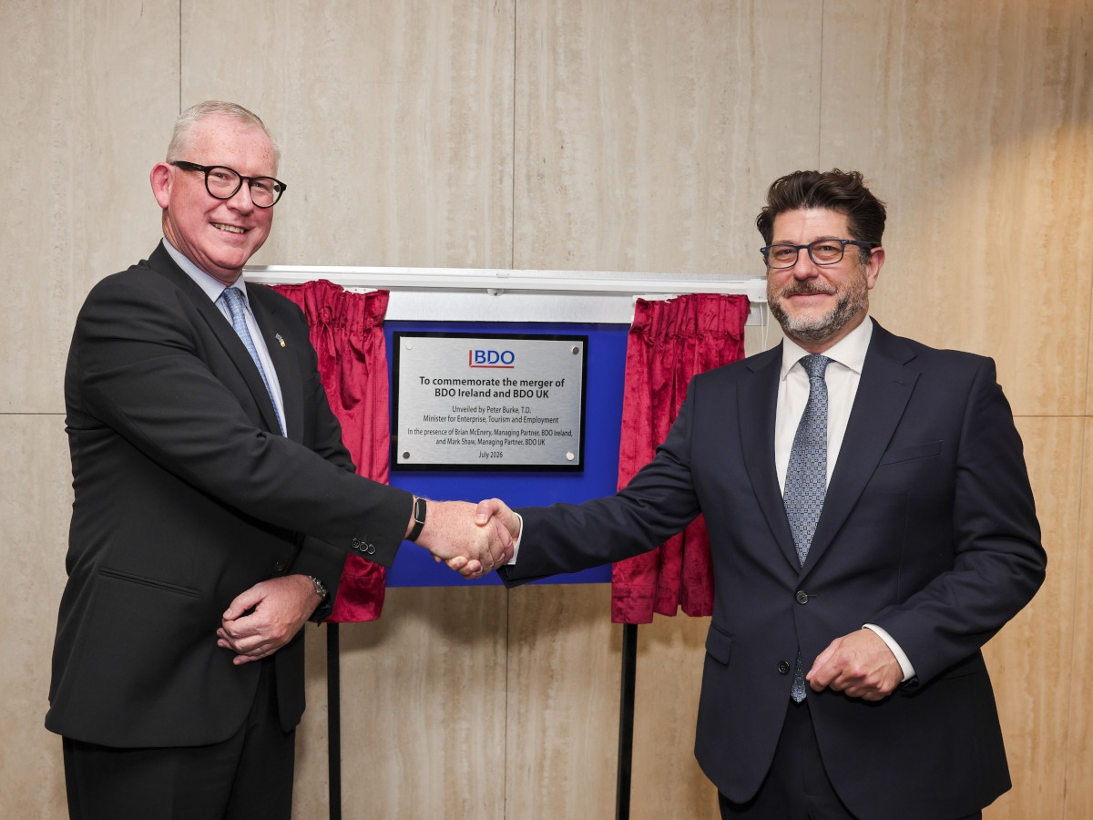 Two white men in suits and ties shake hands in front of a commemorative plaque.