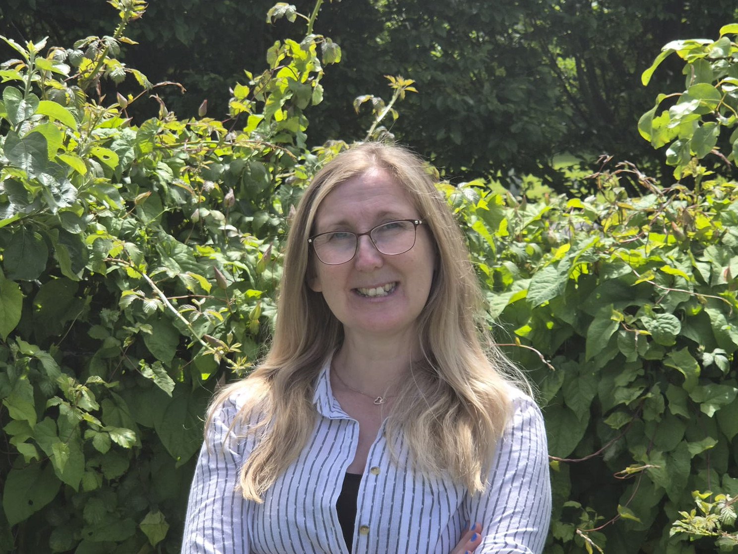 a woman with blonde hair and glasses standing in front of shrubbery.