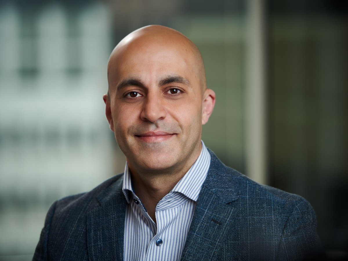 A bald man in a shirt and blazer poses for a headshot in front of a blurred background.