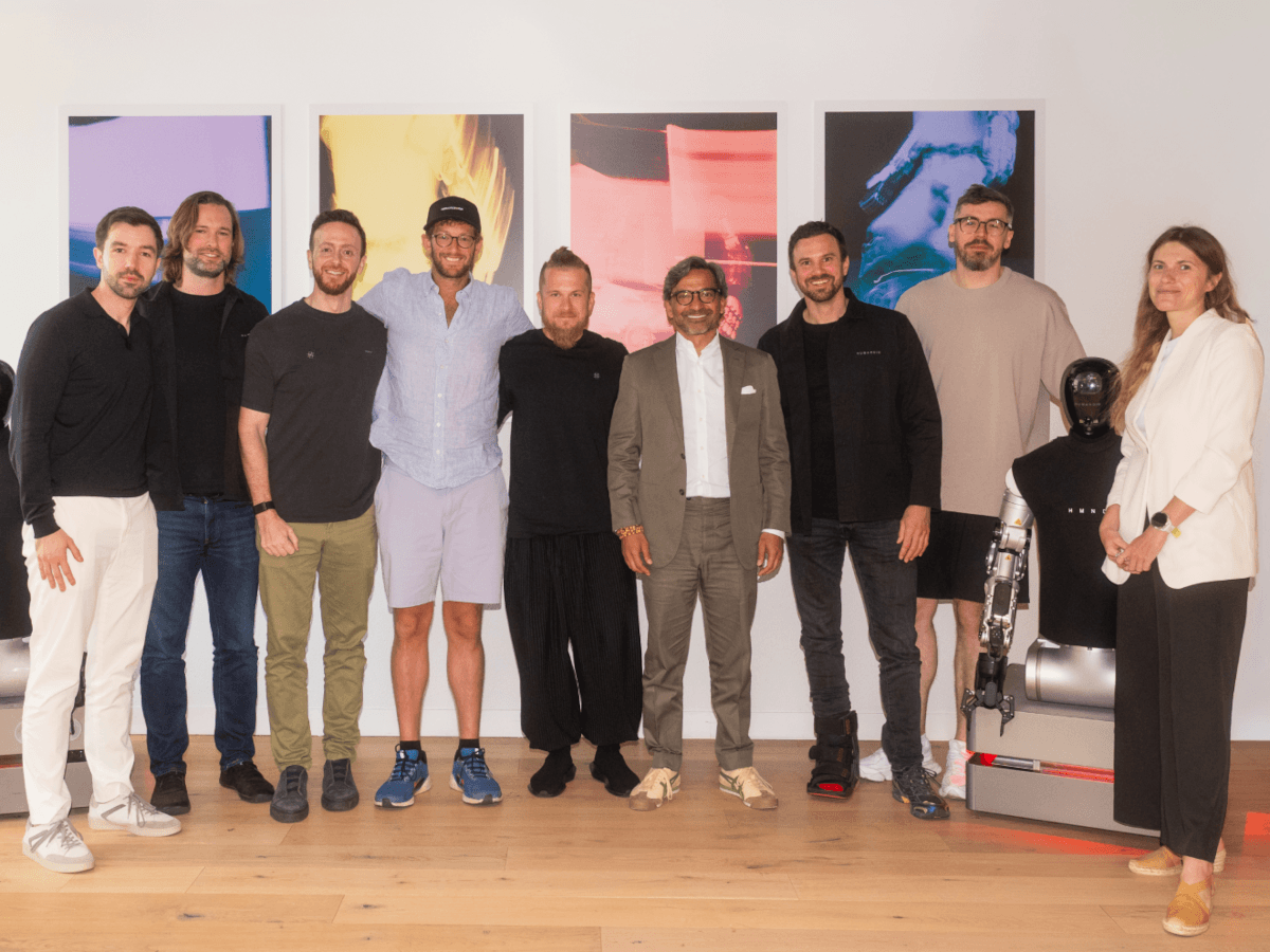 Eight men, one woman and a humanoid robot pose in a line in front of colourful artwork on a white wall.