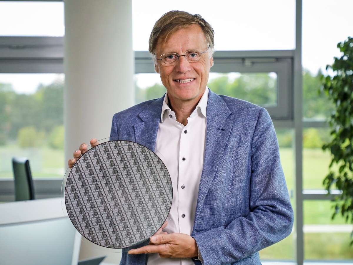 An older man in a blazer and shirt hold a semiconductor wafer in pose for the camera.