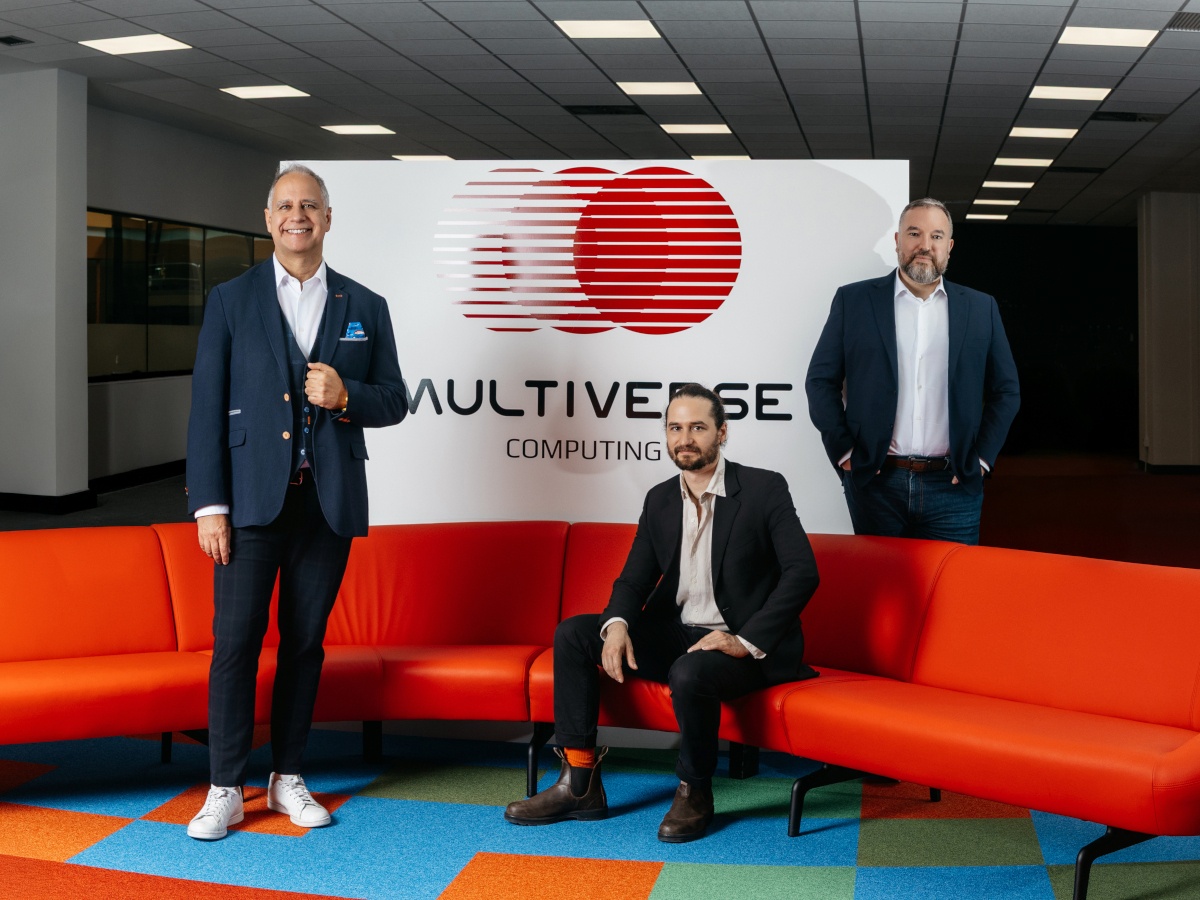 Three men wearing suits smile in front of an orange couch and a wall that displays the Multiverse Computing logo. One of the men is sitting on the couch.
