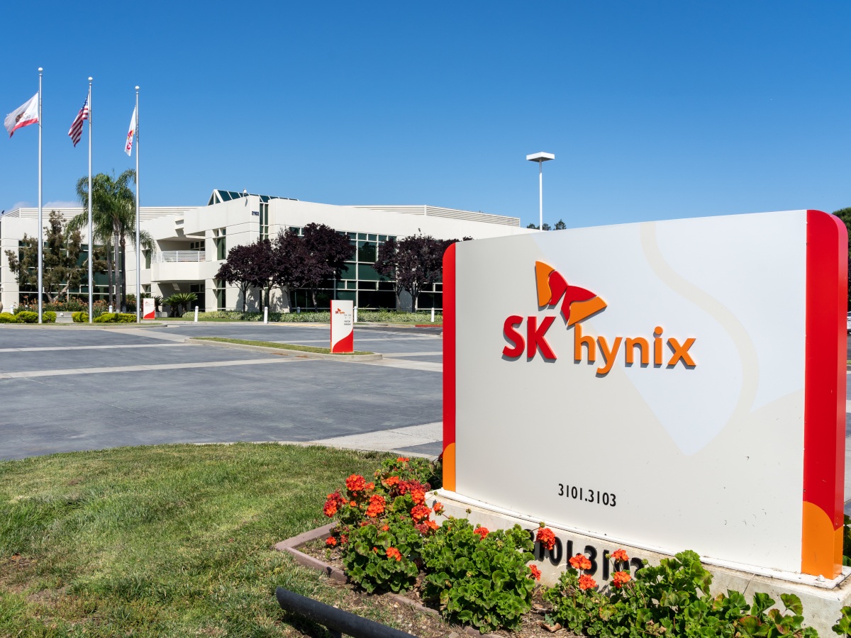 A picture of a sign displaying the SK Hynix logo and name. Behind the sign, is an office complex with flagpoles.