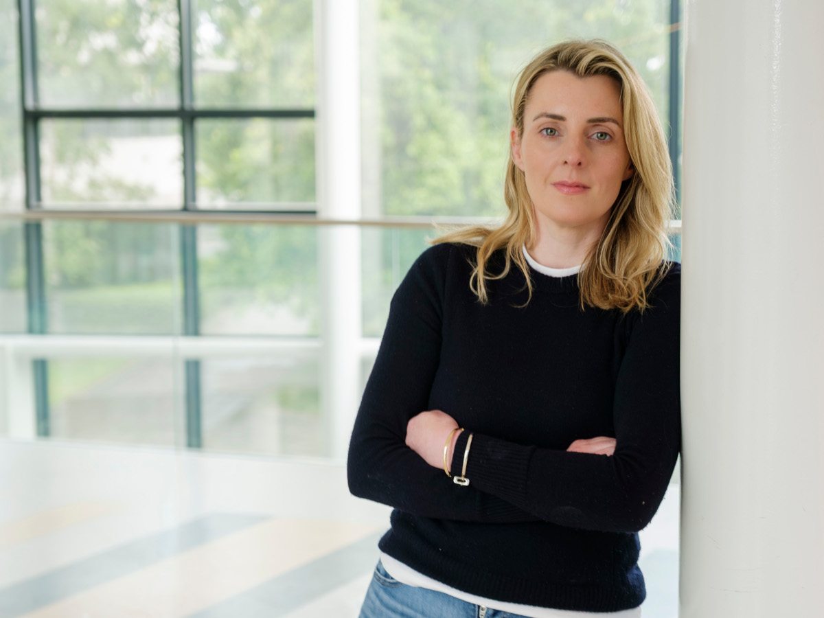 A woman with blonde hair leaning against a wall. She is wearing a black jumper.