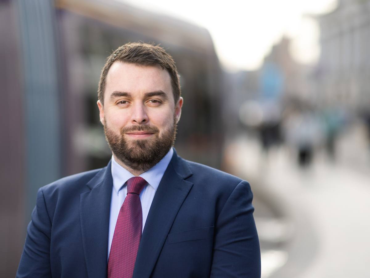 A young white man with short cropped brown hair and beard and a navy suit with a magenta tie.