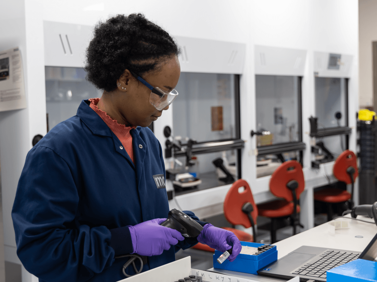 A black woman wearing a lab coat and goggles works at a science lab bench.