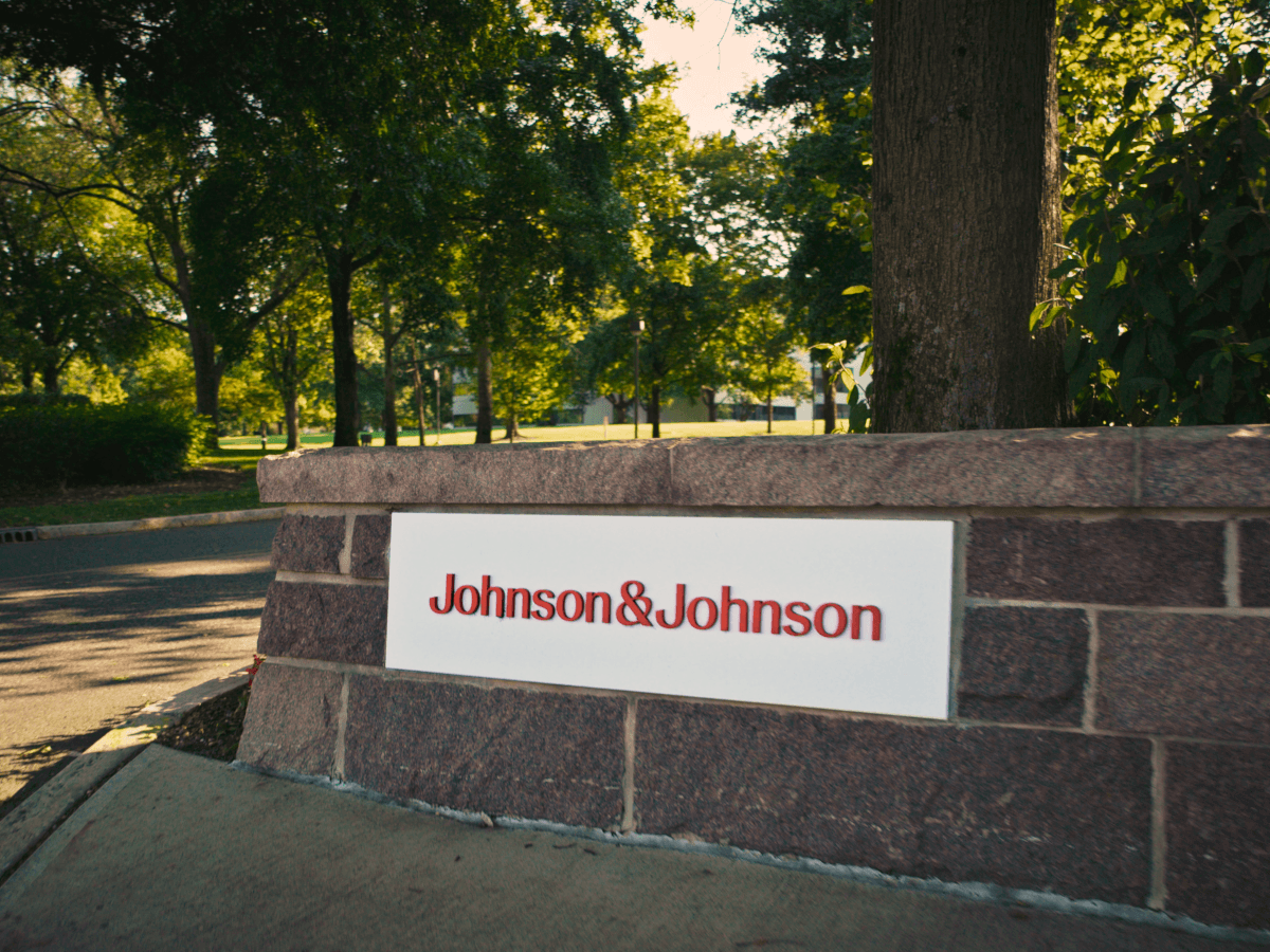 A white and red Johnson & Johnson sign on a brick wall outside a leafy campus.