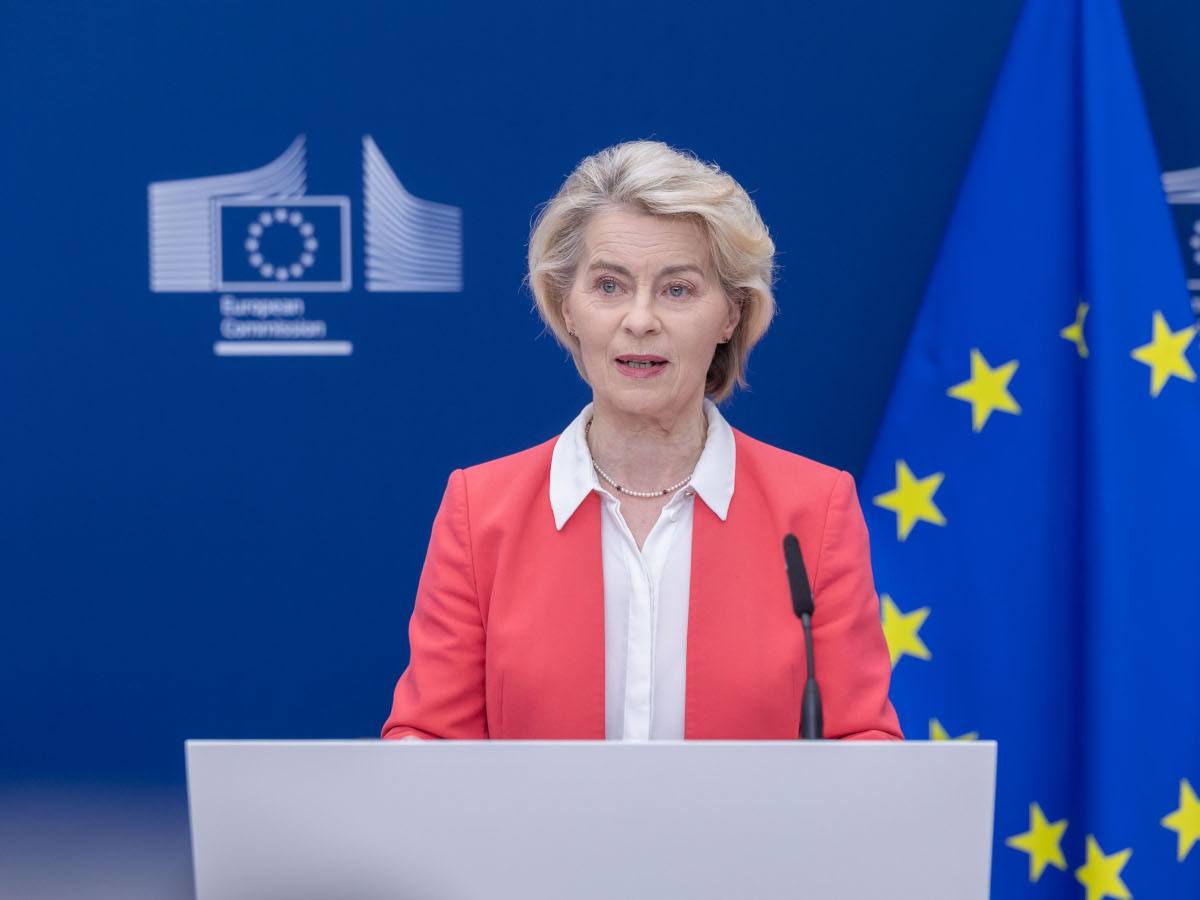 A woman with short blonde hair in a salmon coloured blazer standing on a podium.