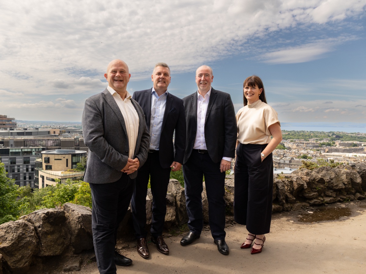 Three men and one woman stand together dressed in business attire smiling, with a scenic view of Edinburgh behind them.