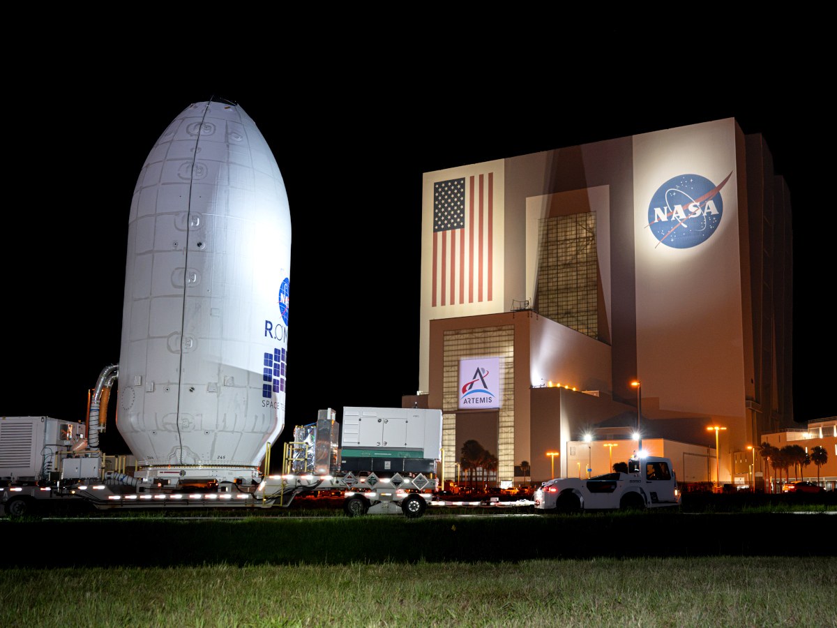 A picture of the NASA Roman Space Telescope inside a massive white capsule outside a NASA facility at night.