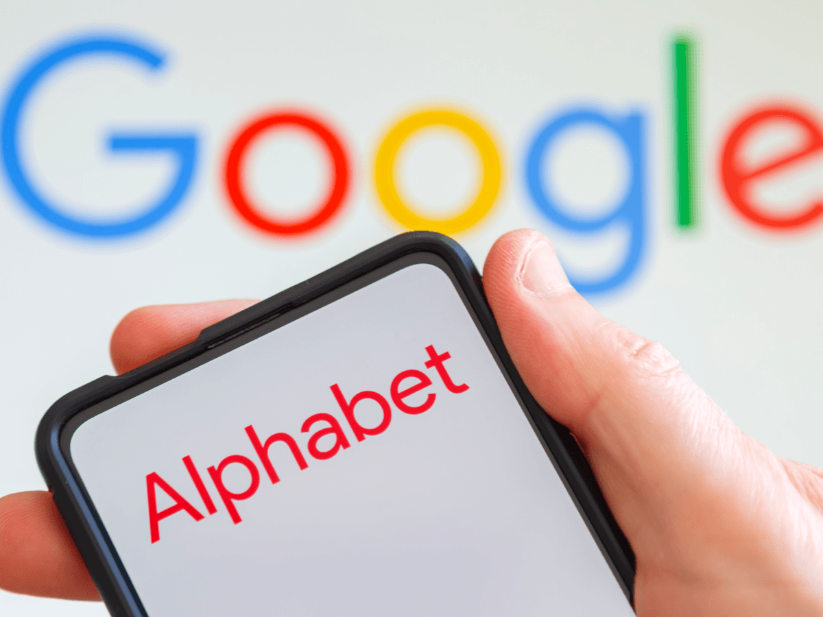 A person holding a phone with Alphabet on it, in front of a blurred Google logo.