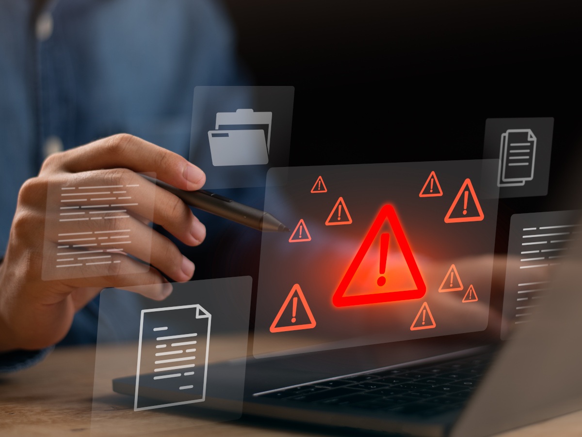 A hand works on a laptop surrounded by cyber warning signals.